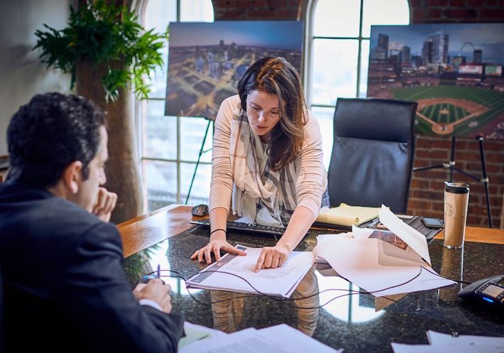 A female executive points out features of a floor plan in a meeting. Behind her are arch windows with city views.