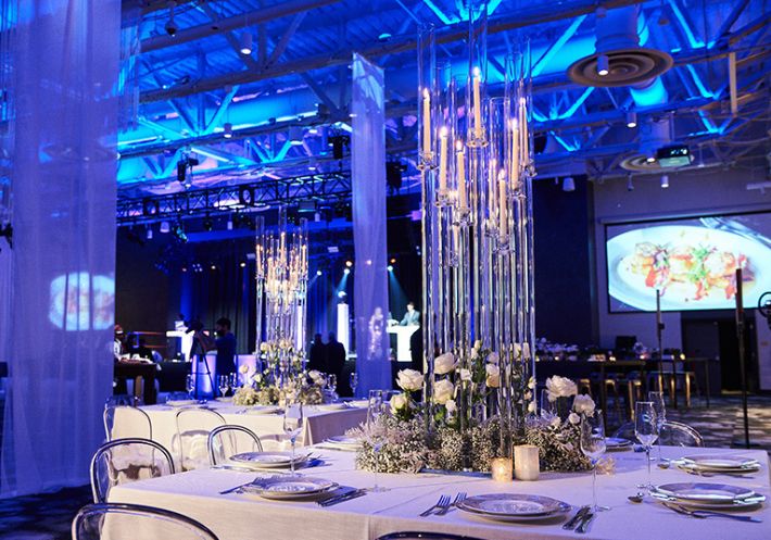 In a blue-lit reception hall, centerpieces with candlesticks in stunning tall glass pillars are encircled by white roses.